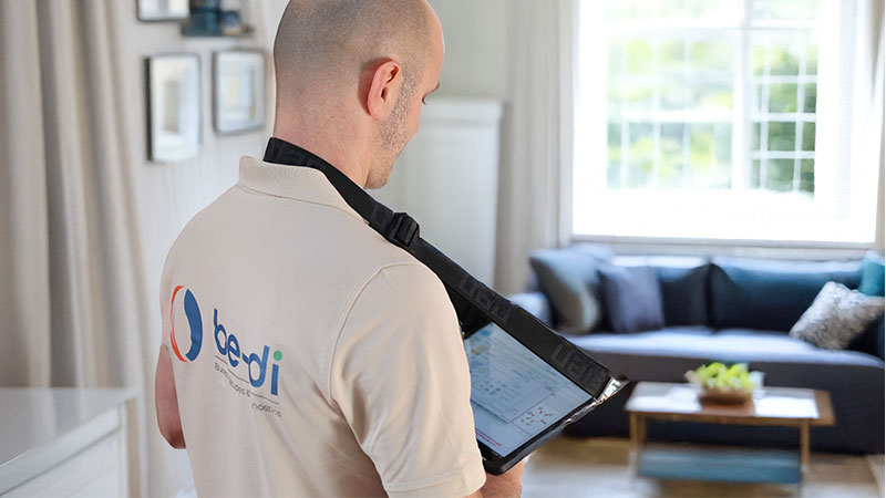 Main tenant une tablette avec une interface de gestion de maison. L'écran affiche un schéma de la maison avec des informations sur la température intérieur, la consommation d'énergie répartie dans les différentes zones de la maison ainsi qu'un graphique de suivi. En arrière-plan, on aperçoit une maison.